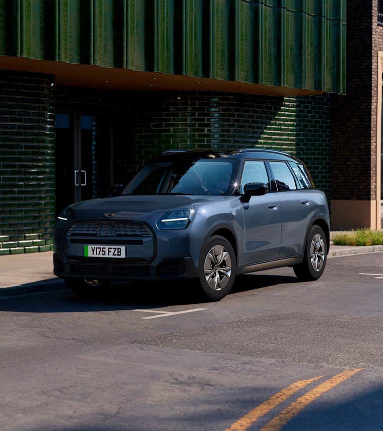 MINI Countryman electric SUV parked on a city street in front of a dark green tiled building, featuring black roof and modern headlights.